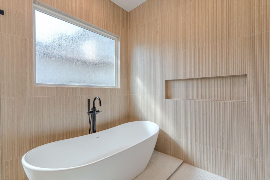 This bathroom features a modern freestanding tub, a sleek black faucet, and textured beige wall tiles. A frosted window provides natural light while maintaining privacy, and a built-in wall niche offers convenient storage.