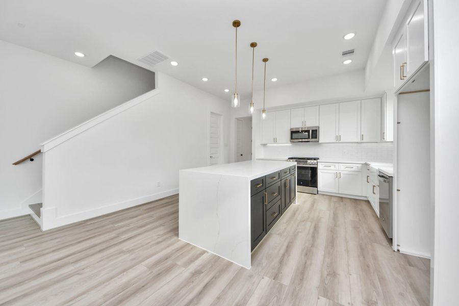This photo showcases a modern kitchen with a spacious, open layout. It features sleek white cabinetry, a large island with dark accents, stainless steel appliances, and elegant pendant lighting. The room has light wood flooring and an adjacent staircase, creating a bright and inviting space.