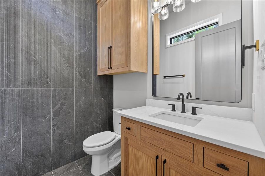 Bathroom featuring dark grey tiled walls and flooring, a light wood-finish vanity with a white countertop and undermount sink, a large rectangular mirror, and an overhead light fixture with glass shades Bathroom featuring dark grey tiled walls and flooring, a light wood-finish vanity with a white countertop and undermount sink, a large rectangular mirror, and an overhead light fixture with glass shades