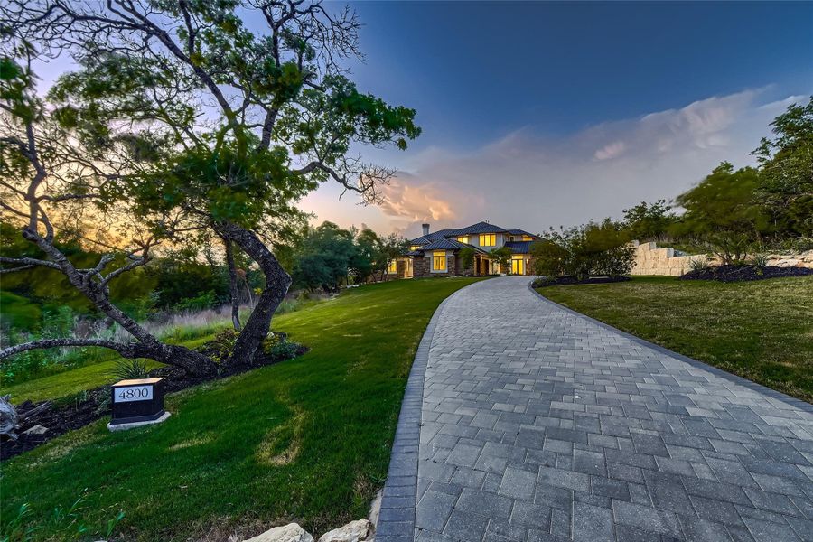 The property features a long, paved driveway, an expansive lawn, and mature trees, leading to a stately home with illuminated windows