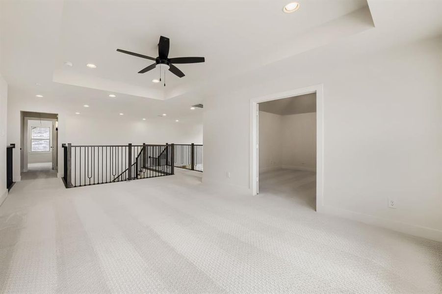 Spacious carpeted landing featuring a tray ceiling with recessed lighting and a ceiling fan, wrought iron balusters, and a doorway opening to an interior room Spacious carpeted landing featuring a tray ceiling with recessed lighting and a ceiling fan, wrought iron balusters, and a doorway opening to an interior room
