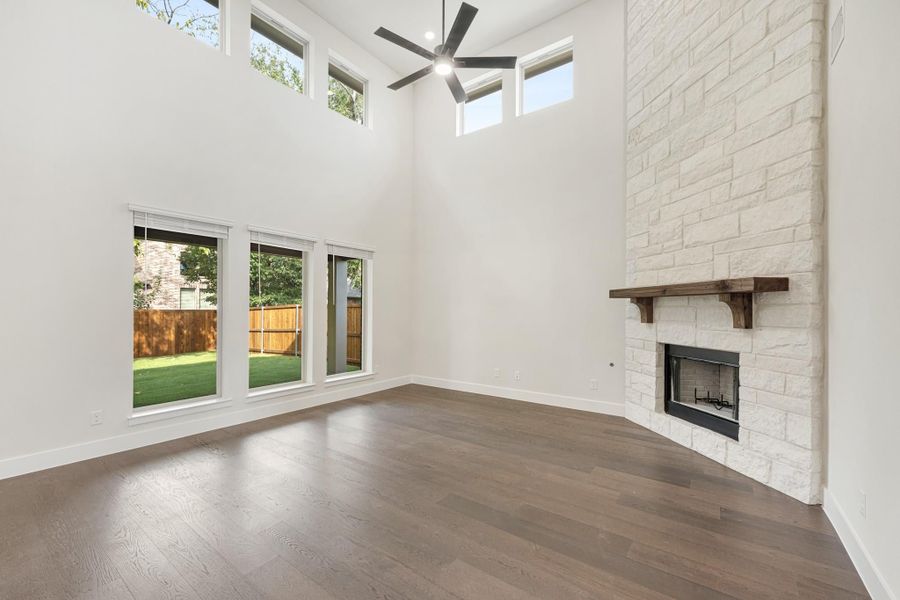 Open living room with two-story ceilings, stone fireplace, hardwood floors, and large windows overlooking backyard