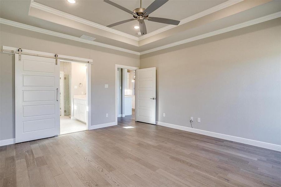 Unfurnished bedroom featuring light hardwood / wood-style flooring, ceiling fan, connected bathroom, a barn door, and a raised ceiling