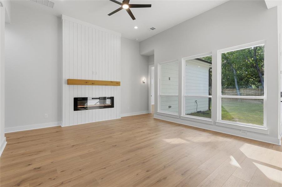 Spacious room featuring light wood-finish flooring, a contemporary fireplace with a wood mantle and shiplap surround, multiple tall windows, a high ceiling, and a ceiling fan with integrated lighting Spacious room featuring light wood-finish flooring, a contemporary fireplace with a wood mantle and shiplap surround, multiple tall windows, a high ceiling, and a ceiling fan with integrated lighting