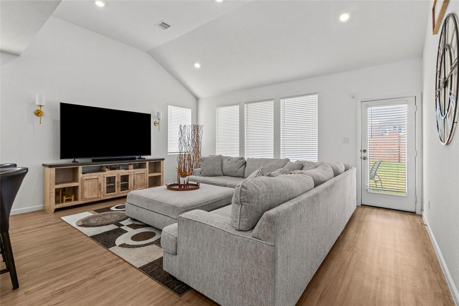 Bright and inviting living area with vaulted ceilings features a spacious sectional sofa, large windows with blinds for natural light, and a modern entertainment setup. A door leads to a backyard space, enhancing indoor-outdoor living. Bright and inviting living area with vaulted ceilings features a spacious sectional sofa, large windows with blinds for natural light, and a modern entertainment setup. A door leads to a backyard space, enhancing indoor-outdoor living.