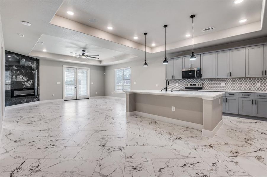 Kitchen with a tray ceiling, an island with sink, gray cabinetry, decorative light fixtures, and light marble finish flooring