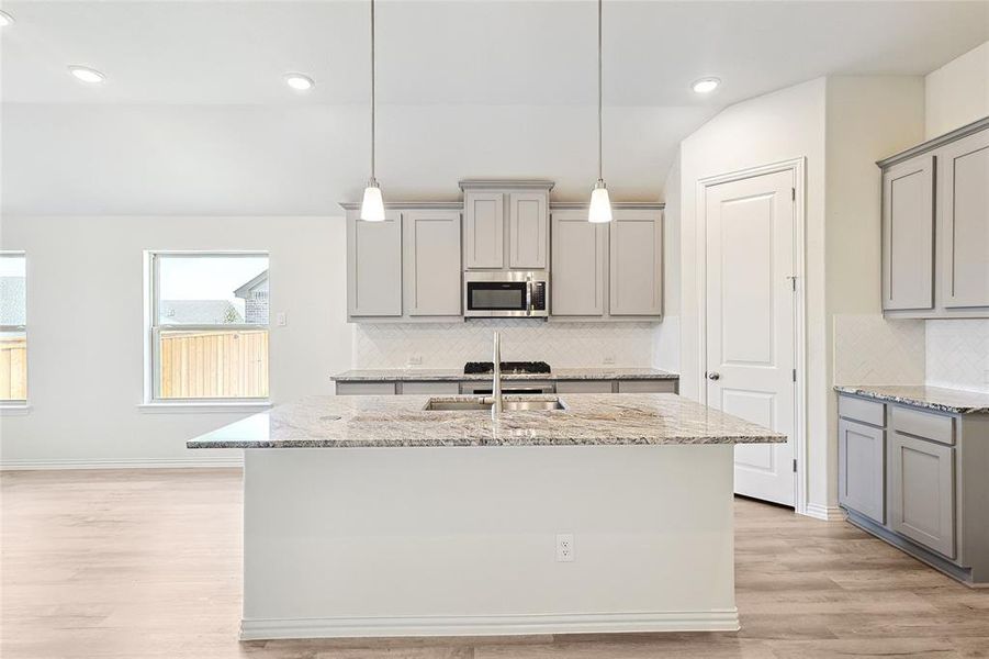 Kitchen featuring hanging light fixtures, decorative backsplash, light hardwood / wood-style floors, an island with sink, and light stone countertops