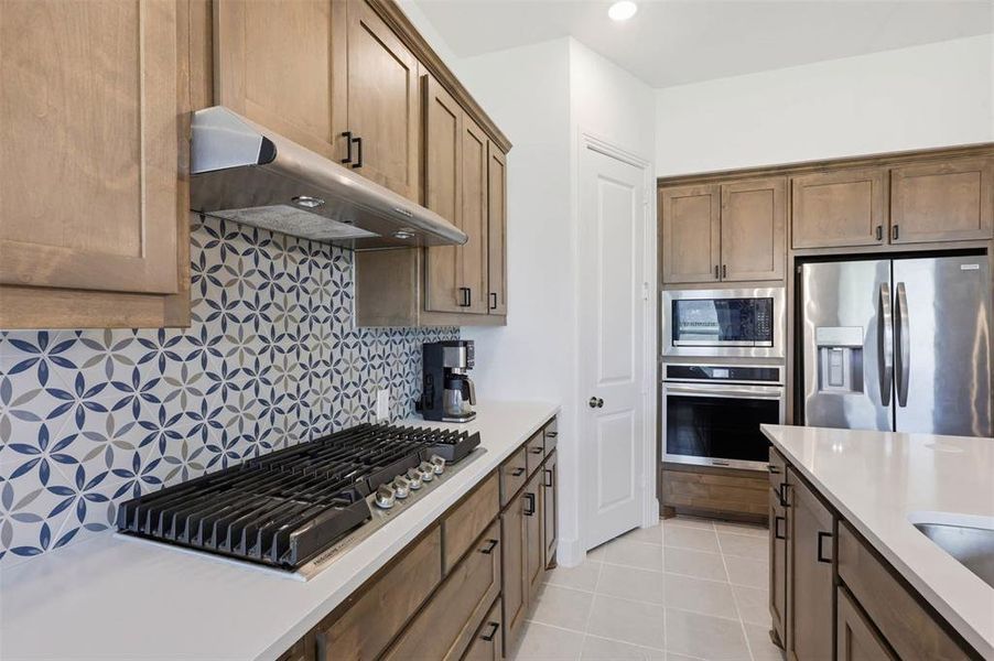 Modern kitchen featuring a patterned tile backsplash, stainless steel appliances, light wood-finish cabinetry, a built-in gas cooktop, and white countertops