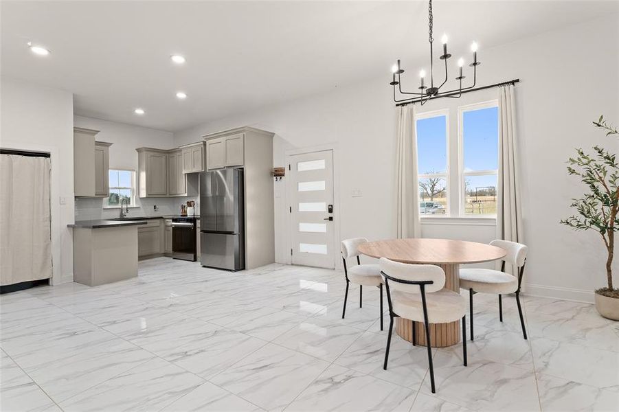Open-concept living space featuring large format marble-look tile flooring, a modern kitchen with grey cabinetry and stainless steel appliances, and a dining area with a contemporary black chandelier Open-concept living space featuring large format marble-look tile flooring, a modern kitchen with grey cabinetry and stainless steel appliances, and a dining area with a contemporary black chandelier