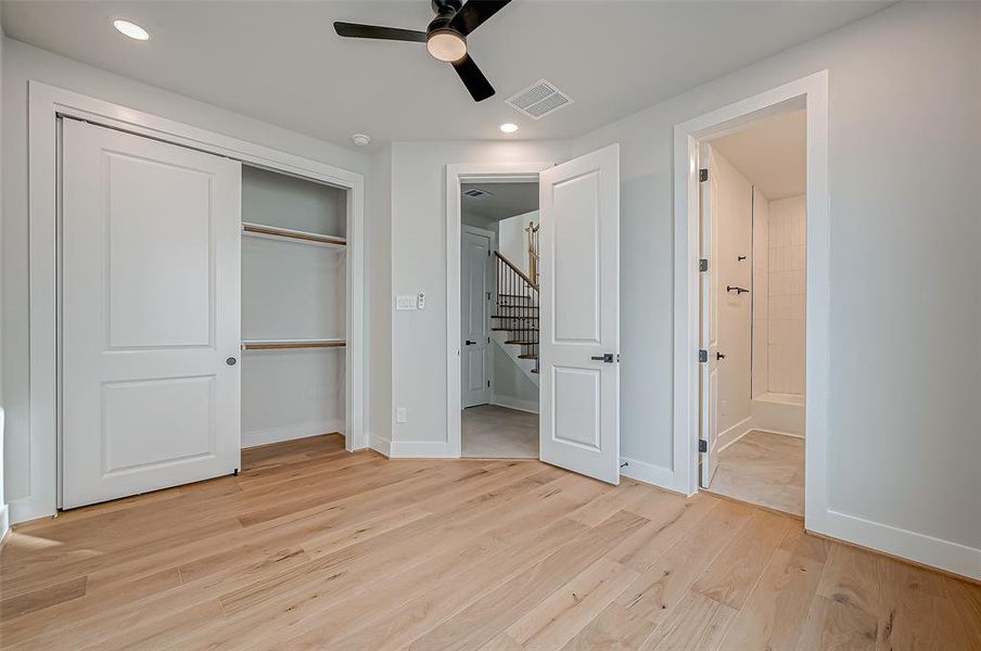 This is a bright, modern bedroom on the first floor with light wood flooring and neutral walls. It features a ceiling fan, a spacious closet with sliding doors, and an en-suite bathroom. The room has recessed lighting and an open doorway leading to a staircase.