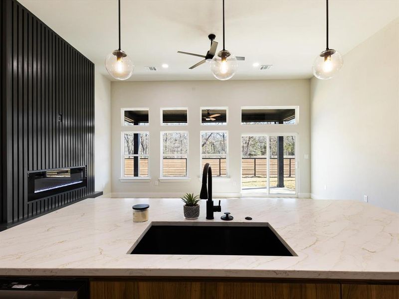 Kitchen with light stone countertops, hanging light fixtures, dishwasher, an island with sink, and a glass covered fireplace