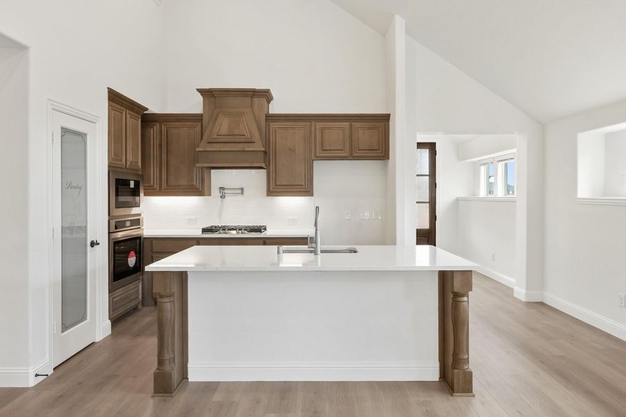 Kitchen with brown wood cabinets, custom range hood, white island with sink, and vaulted ceiling Kitchen with brown wood cabinets, custom range hood, white island with sink, and vaulted ceiling