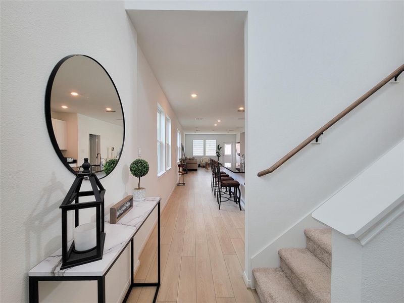 Entryway with modern decor, featuring a large round mirror and a sleek console table. The space leads into an open-plan living area with wooden flooring, ample natural light, and a glimpse of the dining and living rooms. A carpeted staircase is visible on the right.