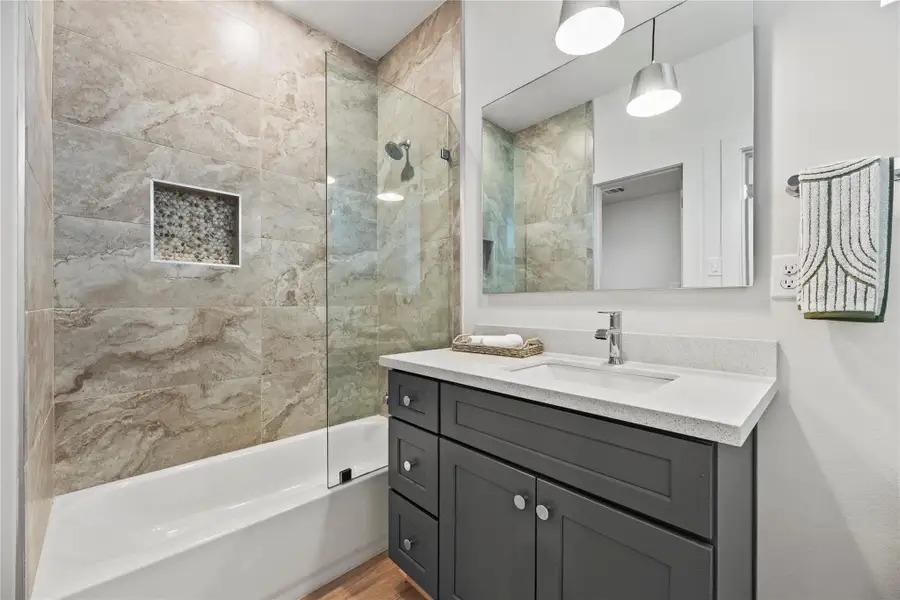 This modern bathroom boasts a sleek vanity with a quartz countertop, a large mirror, and stylish stone-tiled shower with a glass partition and built-in nook. Bright pendant lights enhance the contemporary feel.