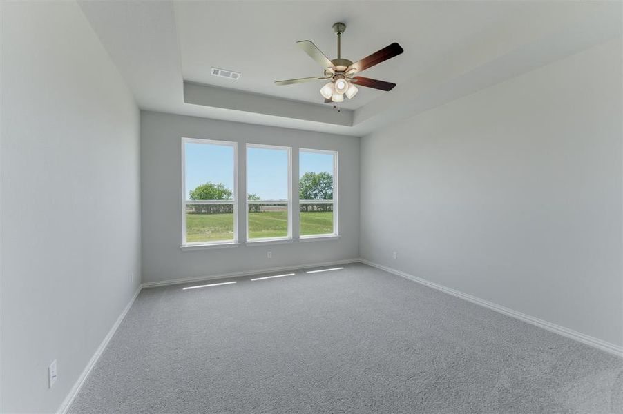 Spacious room featuring three large windows, tray ceiling, ceiling fan with integrated lighting, neutral gray wall paint, and gray carpet flooring Spacious room featuring three large windows, tray ceiling, ceiling fan with integrated lighting, neutral gray wall paint, and gray carpet flooring