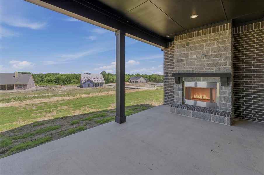 Covered outdoor patio featuring a concrete surface, a built-in fireplace with a stone surround and wood mantel, recessed lighting, and dark brick exterior