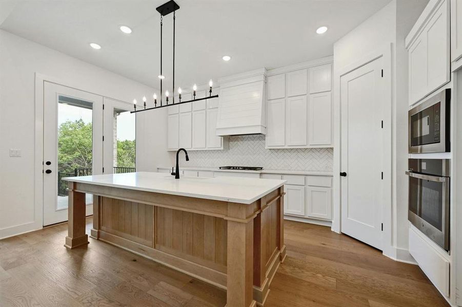 Kitchen featuring backsplash, a center island with sink, pendant lighting, light wood-type flooring, and white cabinetry Kitchen featuring backsplash, a center island with sink, pendant lighting, light wood-type flooring, and white cabinetry