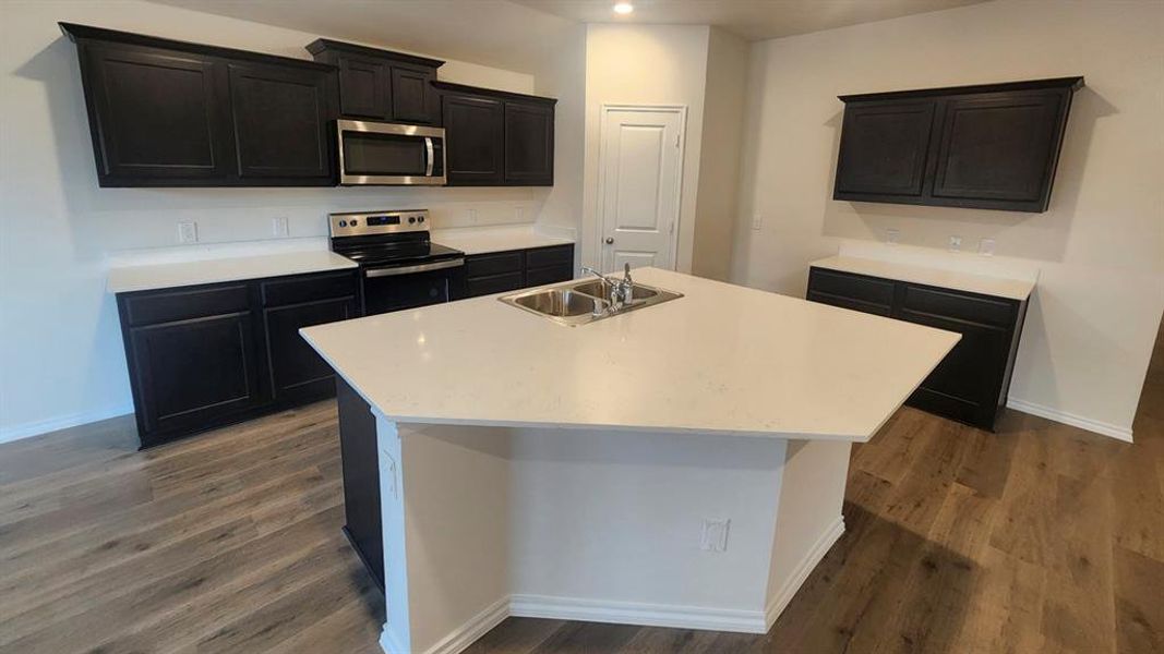 Kitchen featuring a large central island with a double basin sink, white countertops, dark wood-finish cabinetry, stainless steel microwave, and wood-finish flooring