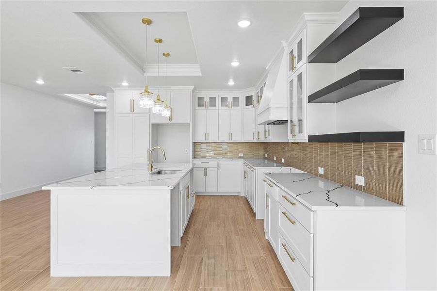 Kitchen with tasteful backsplash, a tray ceiling, light stone counters, glass insert cabinets, and an island with sink Kitchen with tasteful backsplash, a tray ceiling, light stone counters, glass insert cabinets, and an island with sink