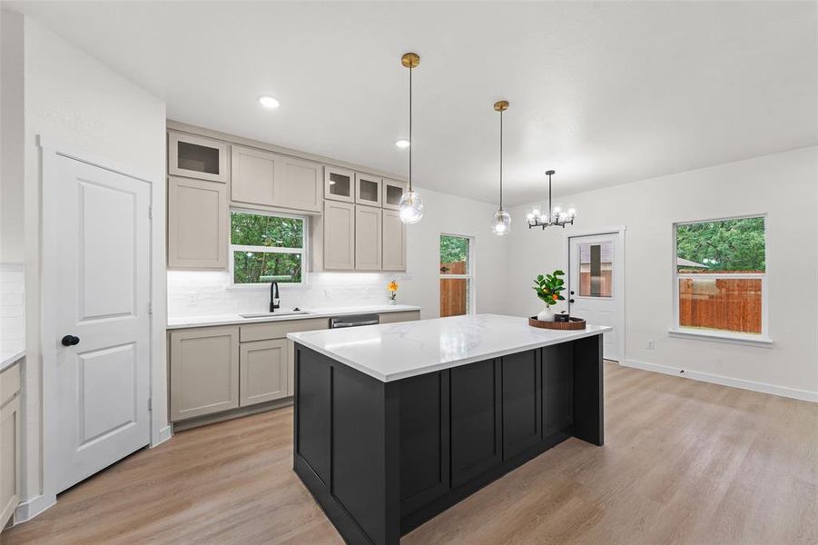Open-concept kitchen featuring an oversized island with a white surface, contrasting dark cabinetry, wood-finish flooring, and pendant lighting Open-concept kitchen featuring an oversized island with a white surface, contrasting dark cabinetry, wood-finish flooring, and pendant lighting