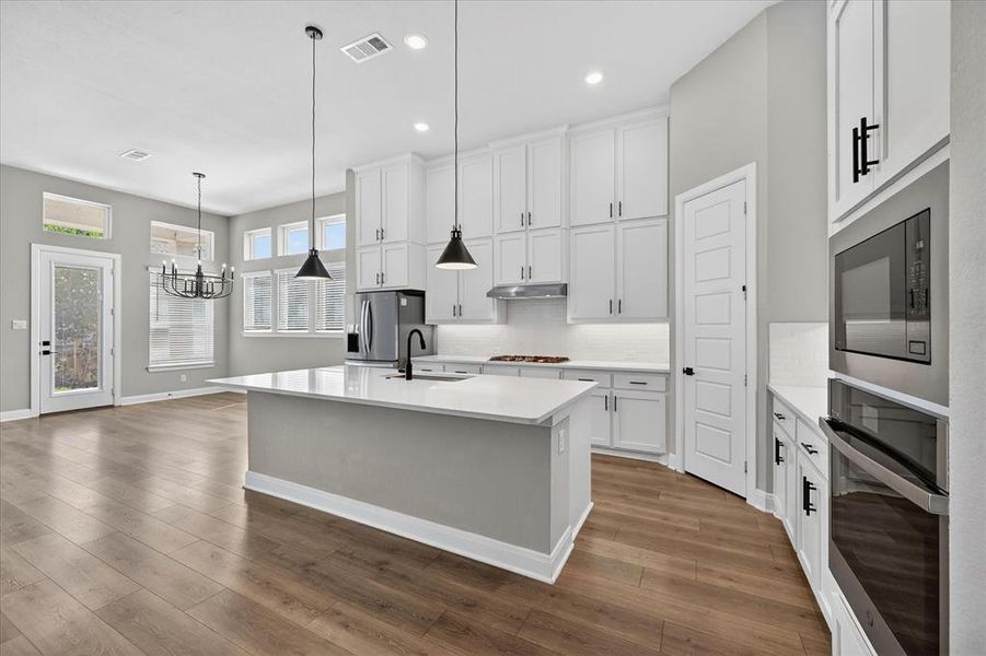 Open-concept kitchen featuring an oversized island with pendant lighting, white shaker cabinetry, stainless steel appliances, and wood-finish flooring Open-concept kitchen featuring an oversized island with pendant lighting, white shaker cabinetry, stainless steel appliances, and wood-finish flooring