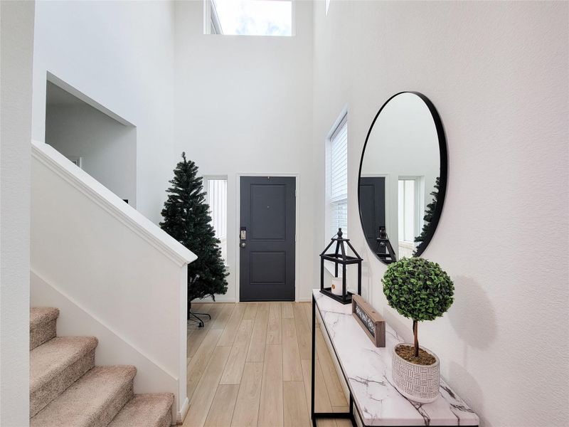 Bright entryway with high ceilings, modern decor, and a staircase. The space features wood-look flooring, a stylish console table with a round mirror, and ample natural light from a large window.