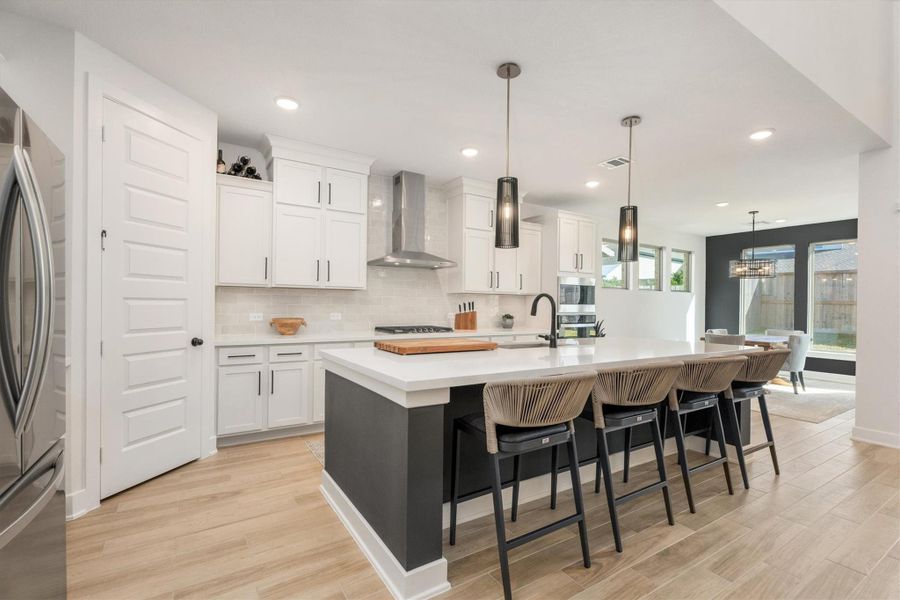 This modern kitchen features sleek white cabinetry, a large island with seating for four, and pendant lighting. It boasts stainless steel appliances, a stylish range hood, and ample counter space, seamlessly flowing into a bright dining area with large windows. Perfect for entertaining and everyday living.