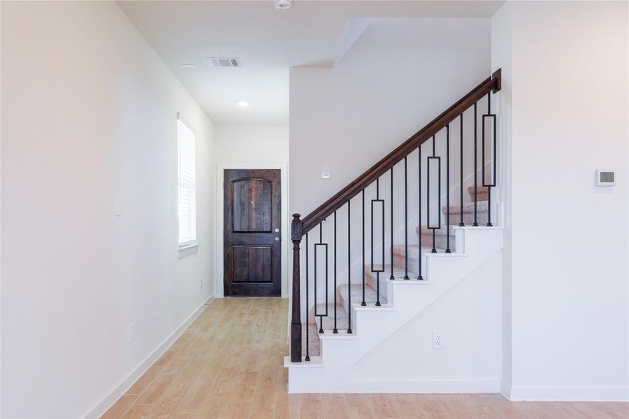 This photo features a bright hallway with light wood flooring and a staircase with elegant dark wood handrails and black metal balusters. A window provides natural light, and a dark wooden door adds a touch of sophistication.