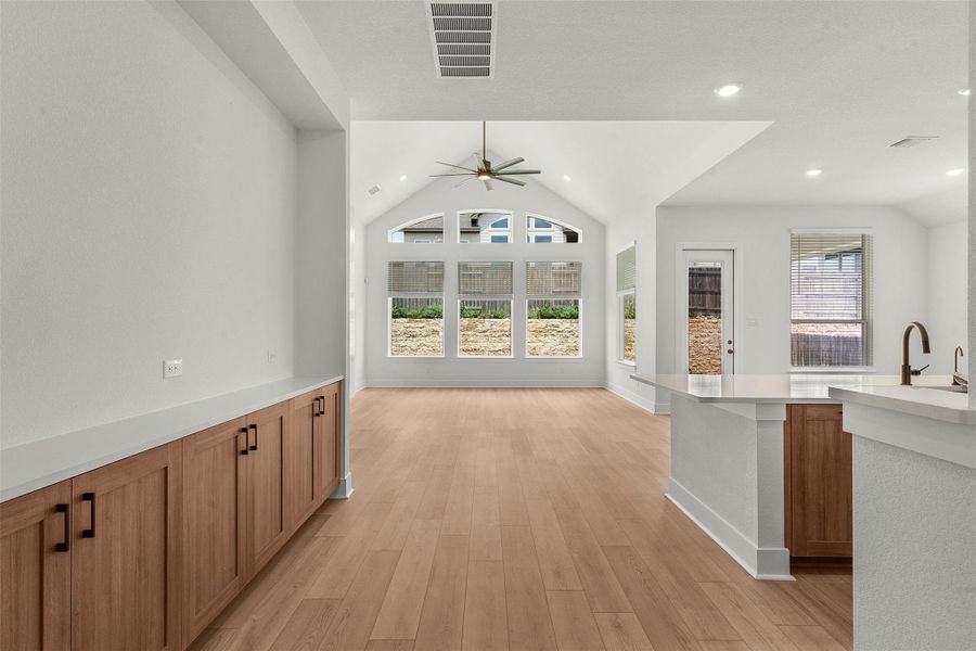 Spacious open-concept living area featuring a vaulted ceiling with a ceiling fan, abundant natural light from large windows, wood-finish flooring, a built-in cabinetry unit with a light-colored countertop, and a kitchen island with a dark faucet Spacious open-concept living area featuring a vaulted ceiling with a ceiling fan, abundant natural light from large windows, wood-finish flooring, a built-in cabinetry unit with a light-colored countertop, and a kitchen island with a dark faucet