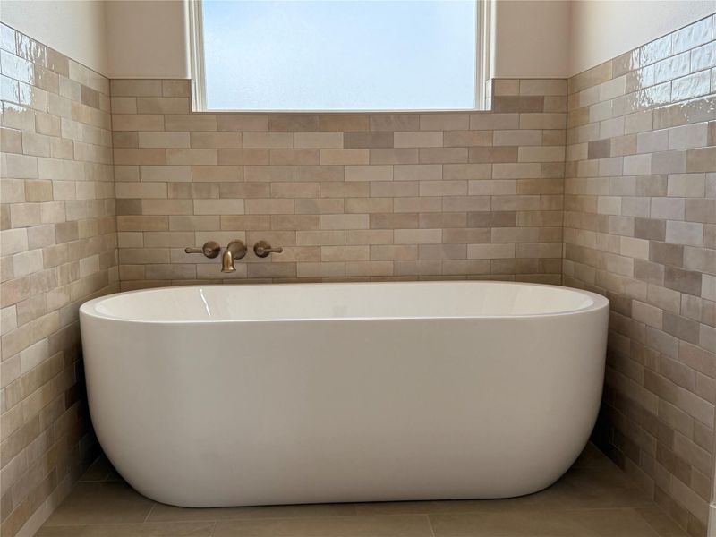 Free standing soaking tub surrounded by an eye catching tile and filled by a gold colored fixture.