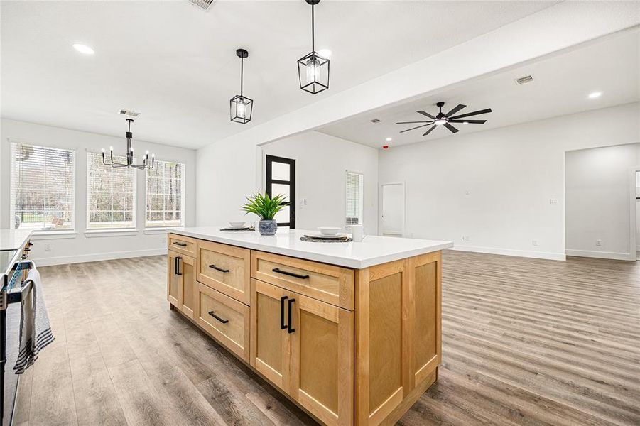 This photo showcases a modern kitchen and open living area with light wood cabinetry, a large island, and contemporary lighting fixtures. The space features sleek appliances, ample counter space, and a seamless transition to the living room with wood-look flooring and a ceiling fan, creating a bright and inviting atmosphere.