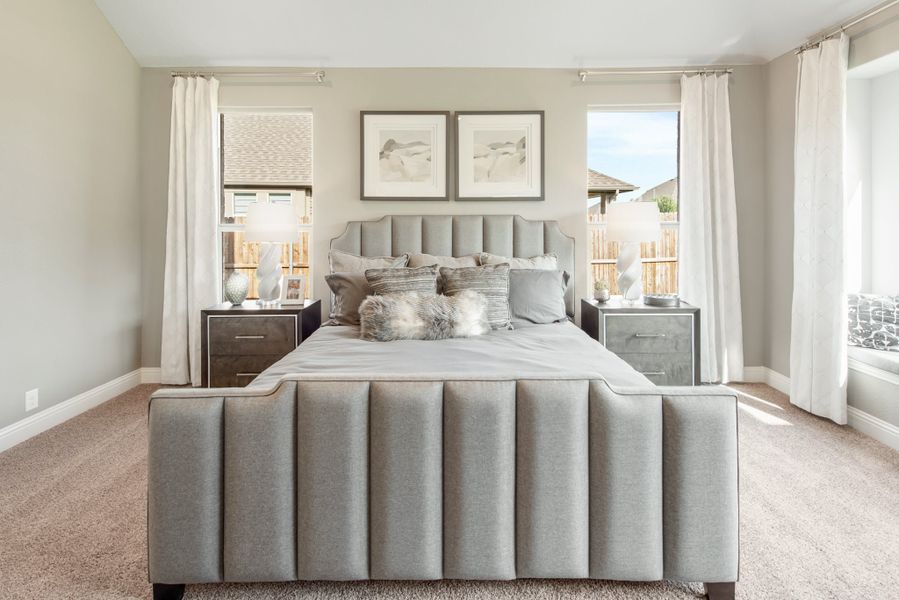 Master bedroom with gray upholstered bed, carpet flooring, two windows with white curtains, and matching nightstands