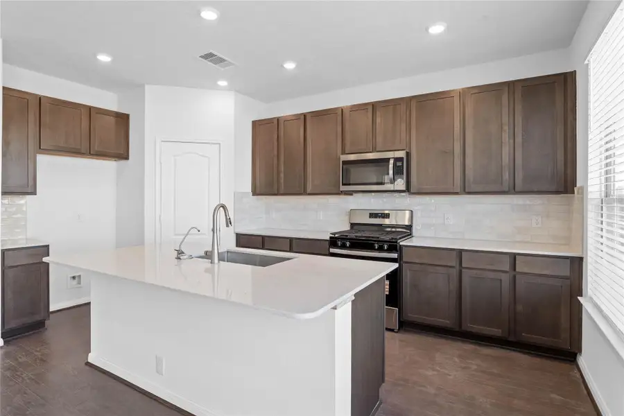This spacious kitchen boasts soaring ceilings, Woodmont Dakota cabinets in a Smolder stain, Silestone Lagoon countertops, stainless steel Whirlpool appliances, a modern Daltile backsplash, recessed and pendant lighting, under-cabinet lighting, an extended breakfast bar countertop, and a pantry—all overlooking the expansive family room.