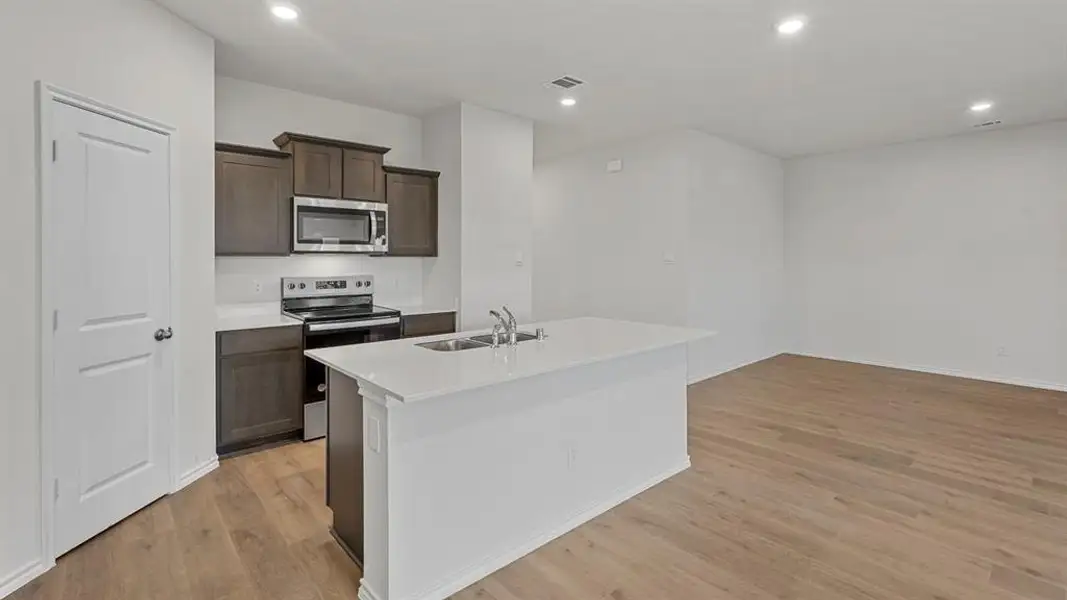 Open-concept living space with wood-finish flooring, a kitchen island with integrated sink, dark wood cabinetry, stainless steel appliances, and recessed lighting