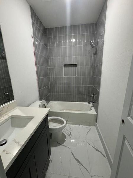 This is a modern bathroom with sleek gray wall tiles and a white bathtub. It features a dark vanity with a white countertop and under-sink storage, complemented by marble-like tile flooring. The space includes a standard toilet and is equipped with a window for natural light and ventilation.