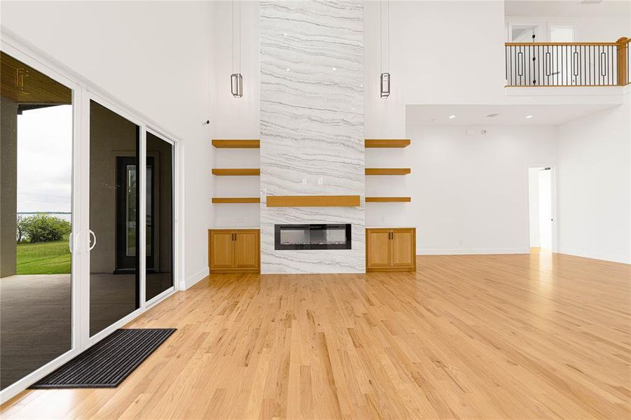 Unfurnished living room with light wood-type flooring, a high ceiling, a premium fireplace, baseboards, and recessed lighting Unfurnished living room with light wood-type flooring, a high ceiling, a premium fireplace, baseboards, and recessed lighting