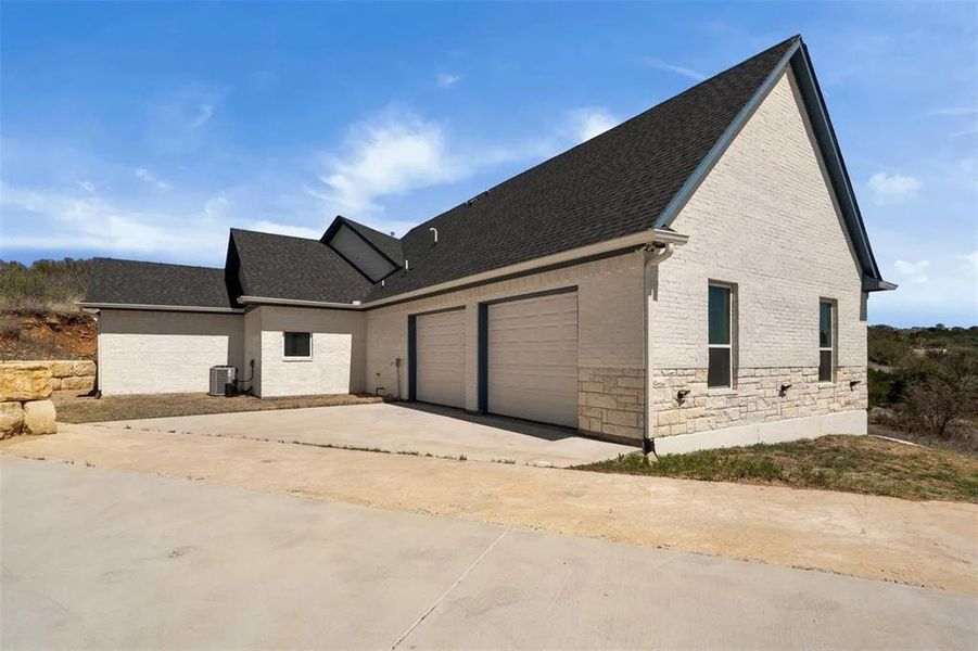 View of front of property featuring a shingled roof, stone siding, driveway, brick siding, and an attached garage