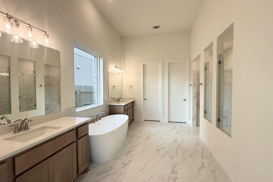 Spacious bathroom featuring a freestanding oval tub, dual vanities with light wood-finish cabinetry and light-toned countertops, and marble-patterned tile flooring