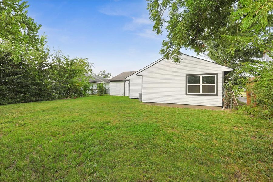 This photo shows a single-story white house with a spacious, well-maintained grassy backyard. The area is bordered by trees, providing natural privacy.