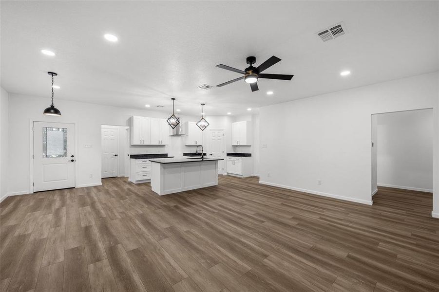 Open-concept living space featuring wood-finish flooring, a modern kitchen island with seating, white shaker cabinetry, dark countertops, and contemporary pendant lighting Open-concept living space featuring wood-finish flooring, a modern kitchen island with seating, white shaker cabinetry, dark countertops, and contemporary pendant lighting