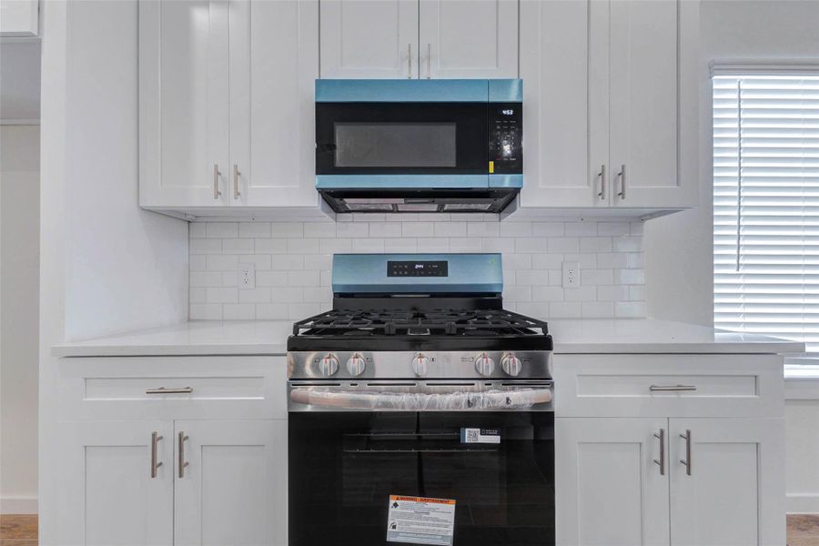 Close-up of the kitchen’s stainless gas range and built-in microwave framed by white shaker cabinetry, sleek hardware, white countertops, and a classic subway tile backsplash. This cooking zone offers a crisp, efficient design with timeless finishes.