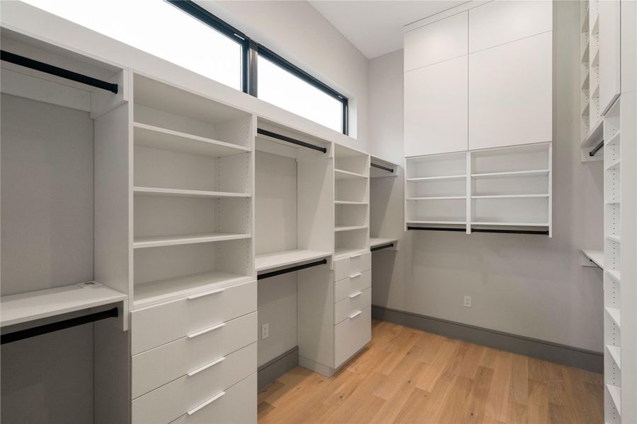 Spacious walk-in closet with ample shelving and drawer space, featuring sleek white cabinetry and natural wood flooring. High windows provide natural light, enhancing the room's modern and organized feel.
