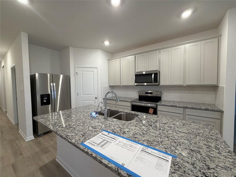 Kitchen with light stone countertops, appliances with stainless steel finishes, tasteful backsplash, a center island with sink, and light wood-style flooring Kitchen with light stone countertops, appliances with stainless steel finishes, tasteful backsplash, a center island with sink, and light wood-style flooring