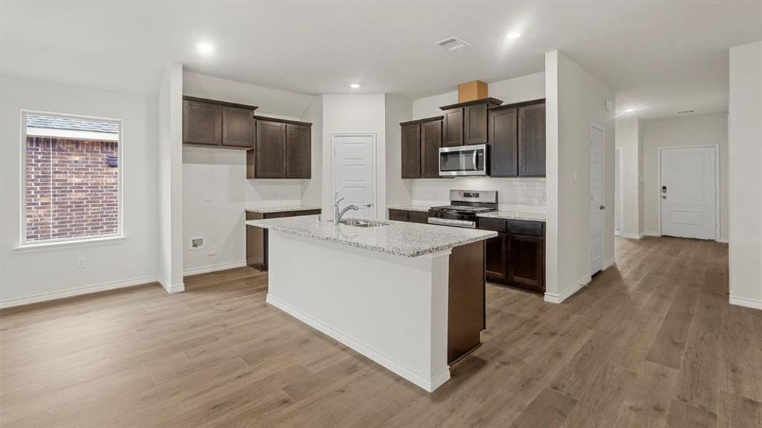 Kitchen with dark brown cabinets, light stone countertops, appliances with stainless steel finishes, an island with sink, and light wood-type flooring Kitchen with dark brown cabinets, light stone countertops, appliances with stainless steel finishes, an island with sink, and light wood-type flooring