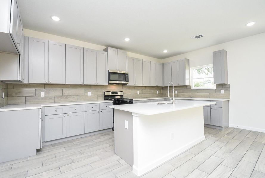This is a modern kitchen featuring gray shaker-style cabinets, stainless steel appliances, a central island with a sink, and light wood-look tile flooring. There's recessed lighting and a window providing natural light. This is a modern kitchen featuring gray shaker-style cabinets, stainless steel appliances, a central island with a sink, and light wood-look tile flooring. There's recessed lighting and a window providing natural light.