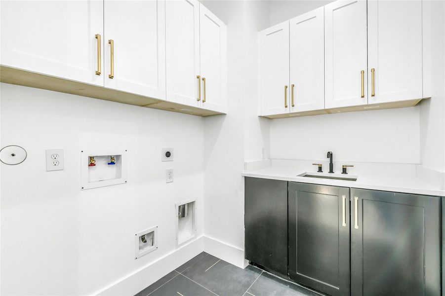 The laundry room combines modern convenience with sleek design. Outfitted with upper and lower custom cabinetry, quartz countertops, and brushed brass hardware, the space offers both durability and sophistication. A deep under-mount utility sink with matte black faucet, full appliance hookups, and oversized tile flooring complete this utility space built for efficiency and refined practicality.