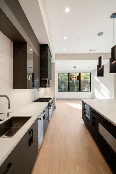 This photo showcases a modern kitchen with sleek dark cabinetry, light countertops, and stainless steel appliances. It features a large island with pendant lighting, an open layout, and ample natural light from a large window.