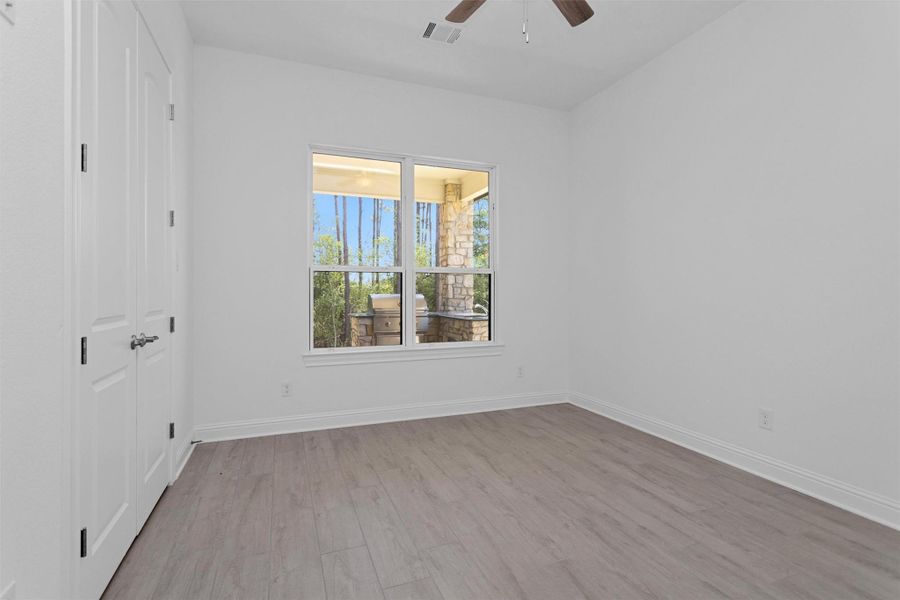 Secondary bedroom features soft neutral wood-look tile flooring, a double-door closet, and a picture window overlooking the outdoor kitchen and lush wooded backdrop.