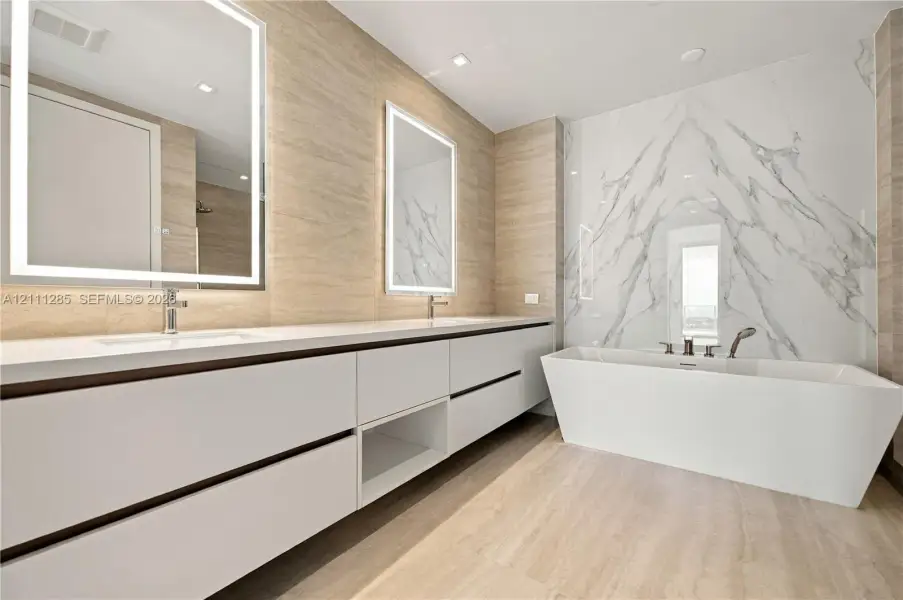 Dual-sink vanity with white cabinetry and countertop, backlit rectangular mirrors, freestanding soaking tub, and large-format tile surround Dual-sink vanity with white cabinetry and countertop, backlit rectangular mirrors, freestanding soaking tub, and large-format tile surround