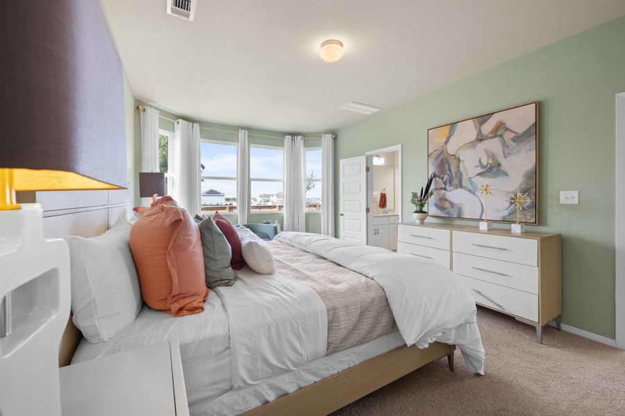 An interior image of the bedroom with green painted walls, brown carpet, a bed with white bedding with colorful pillows, green chairs and wall art.